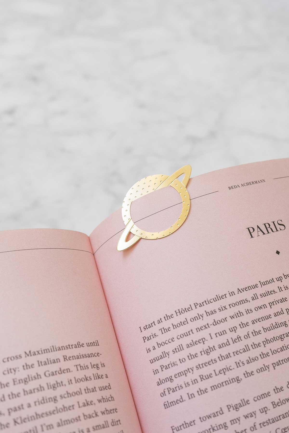 A metal brass Clip by OCTAEVO, gold Saturn-shaped, rests on the open pages of a pink book. 