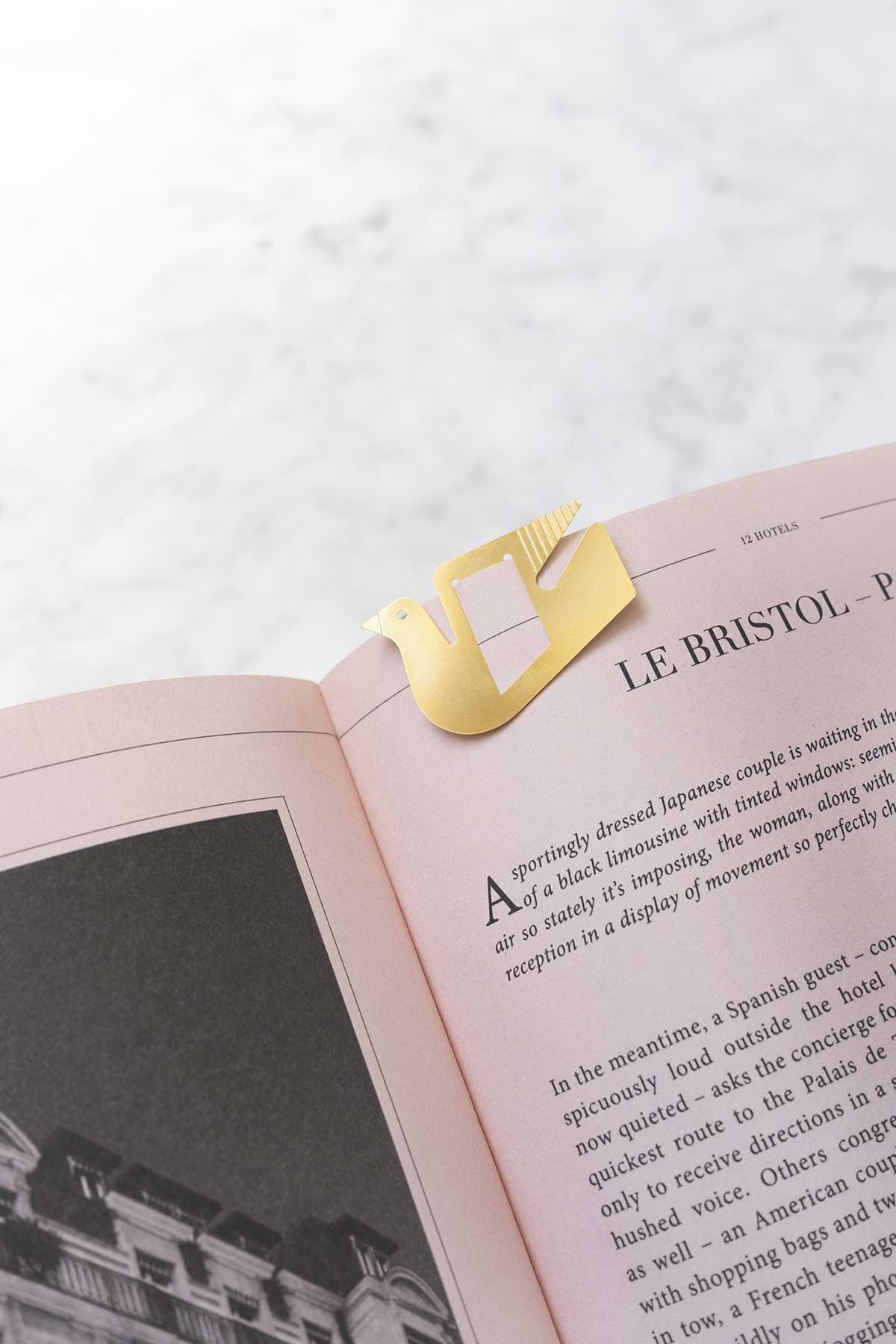 Gold bird-shaped metal brass clip by OCTAEVO clips onto pink-edged pages with Mediterranean charm.