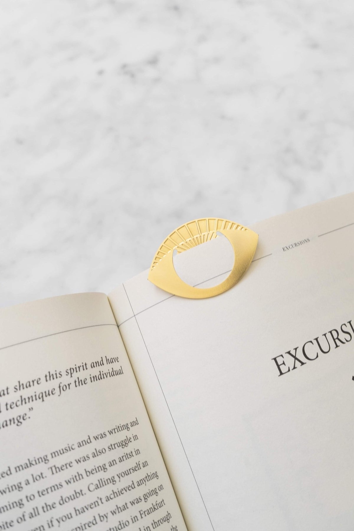 The OCTAEVO Metal Clips Talisman, a gold eye-shaped accessory, decorates an open book on a light marble surface.