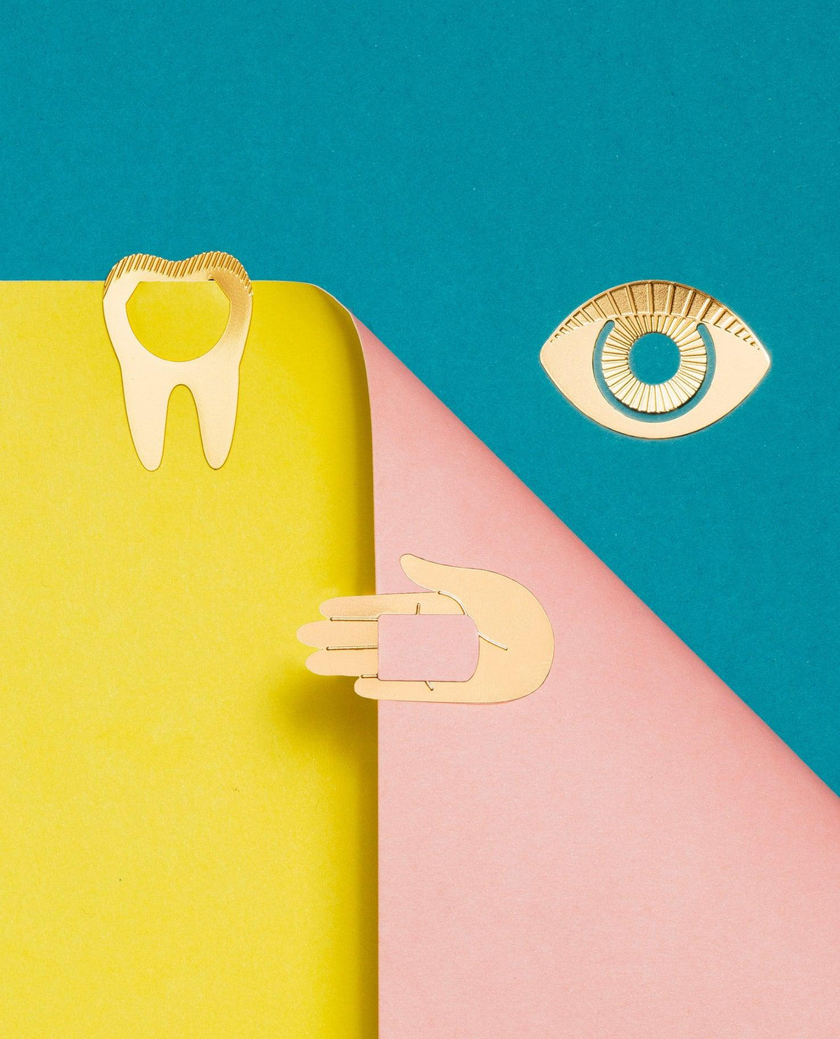 Clips métalliques Talisman OCTAEVO : une dent, un œil et une main dorés à collectionner sur du papier jaune, rose et bleu canard éclatant—des porte-bonheur.