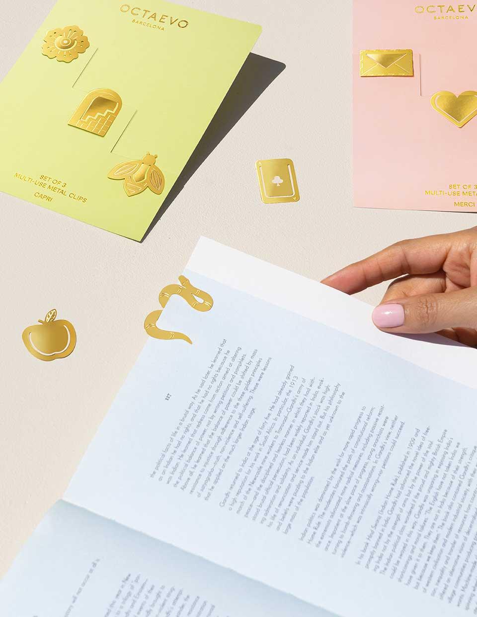 A hand places an OCTAEVO Metal Clips Templo on a blue book, with pastel cards and gold clips nearby.