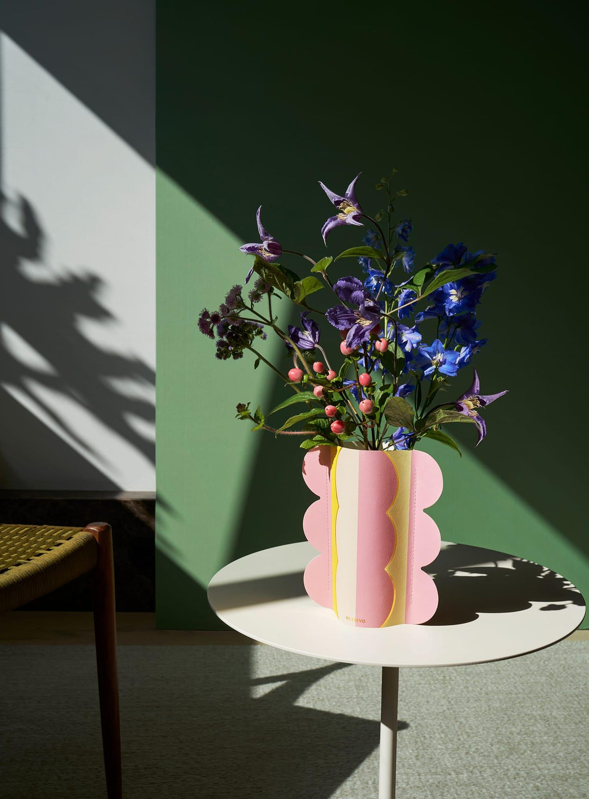 Le mini vase en papier Riviera Wave OCTAEVO garni de fleurs est posé sur une table ronde blanche devant un mur vert.