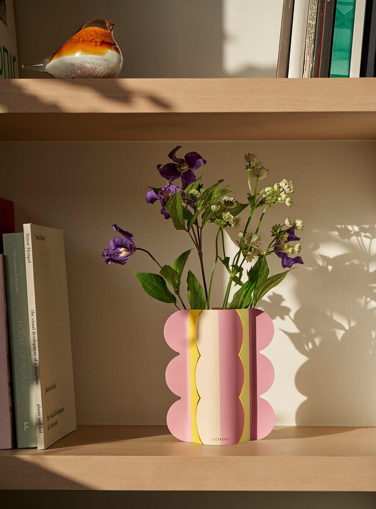 Le mini vase en papier Riviera Wave OCTAEVO apporte un charme méditerranéen avec des fleurs violettes et blanches—idéal pour une étagère à livres.