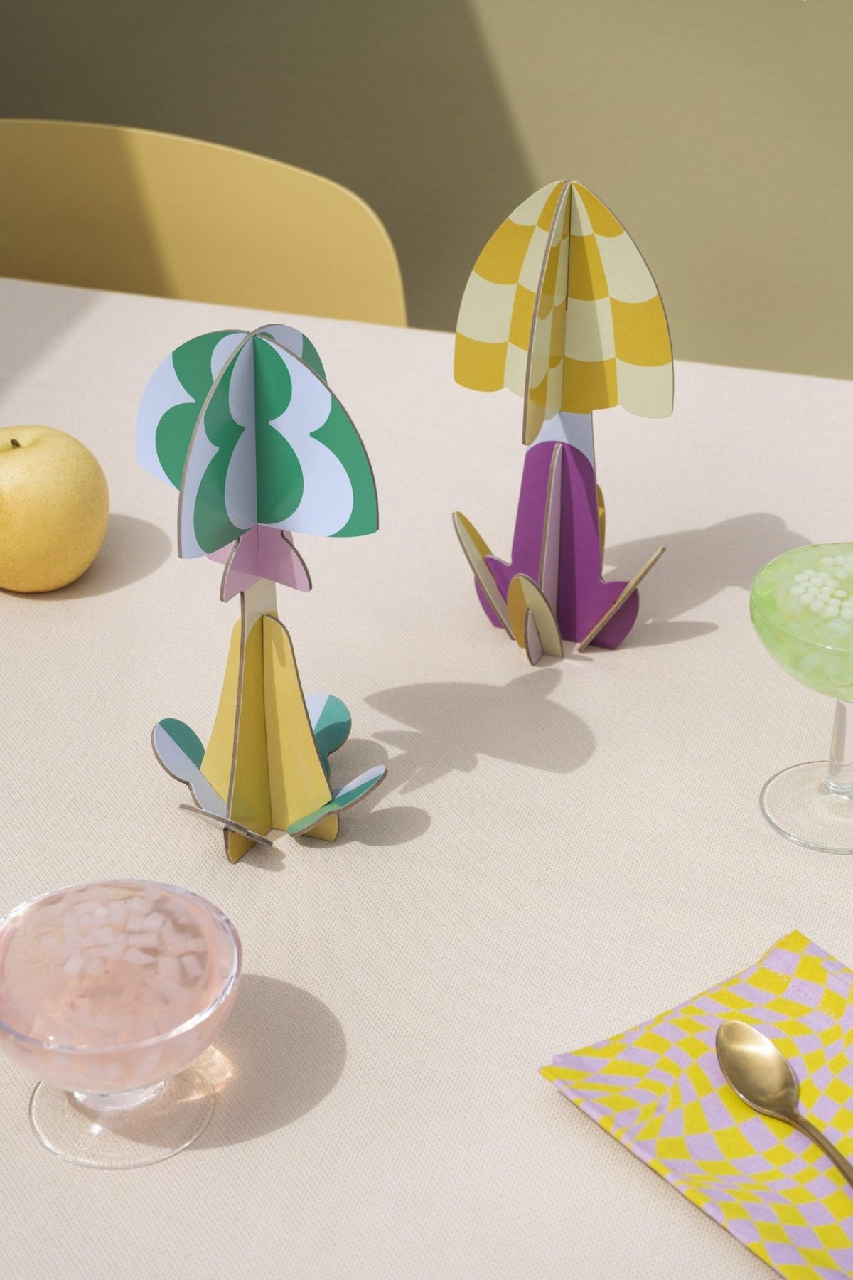 Moss Mushroom Paper Sculpture by OCTAEVO with 3D fruits and drinks on a light table, yellow chair, and playful decor.