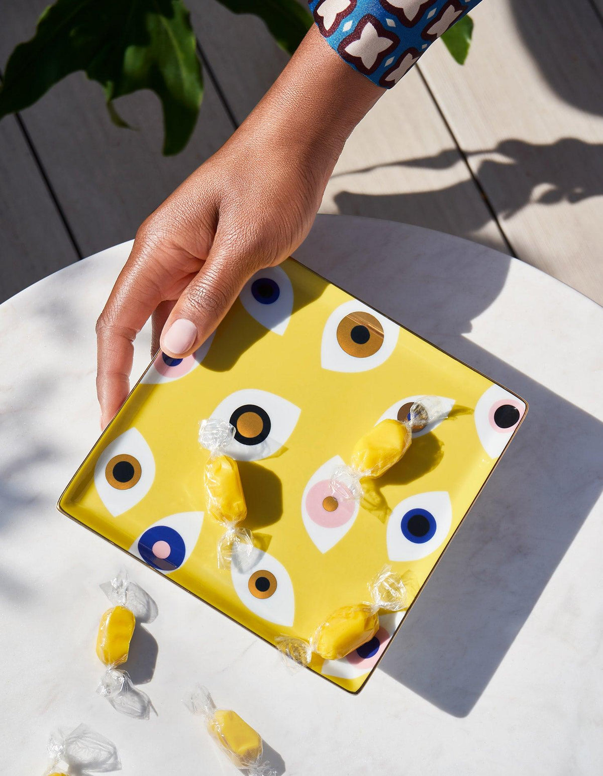 A hand reaches for yellow candies on OCTAEVO Nazar Ceramic Tray, featuring colorful eyes, on a white table.