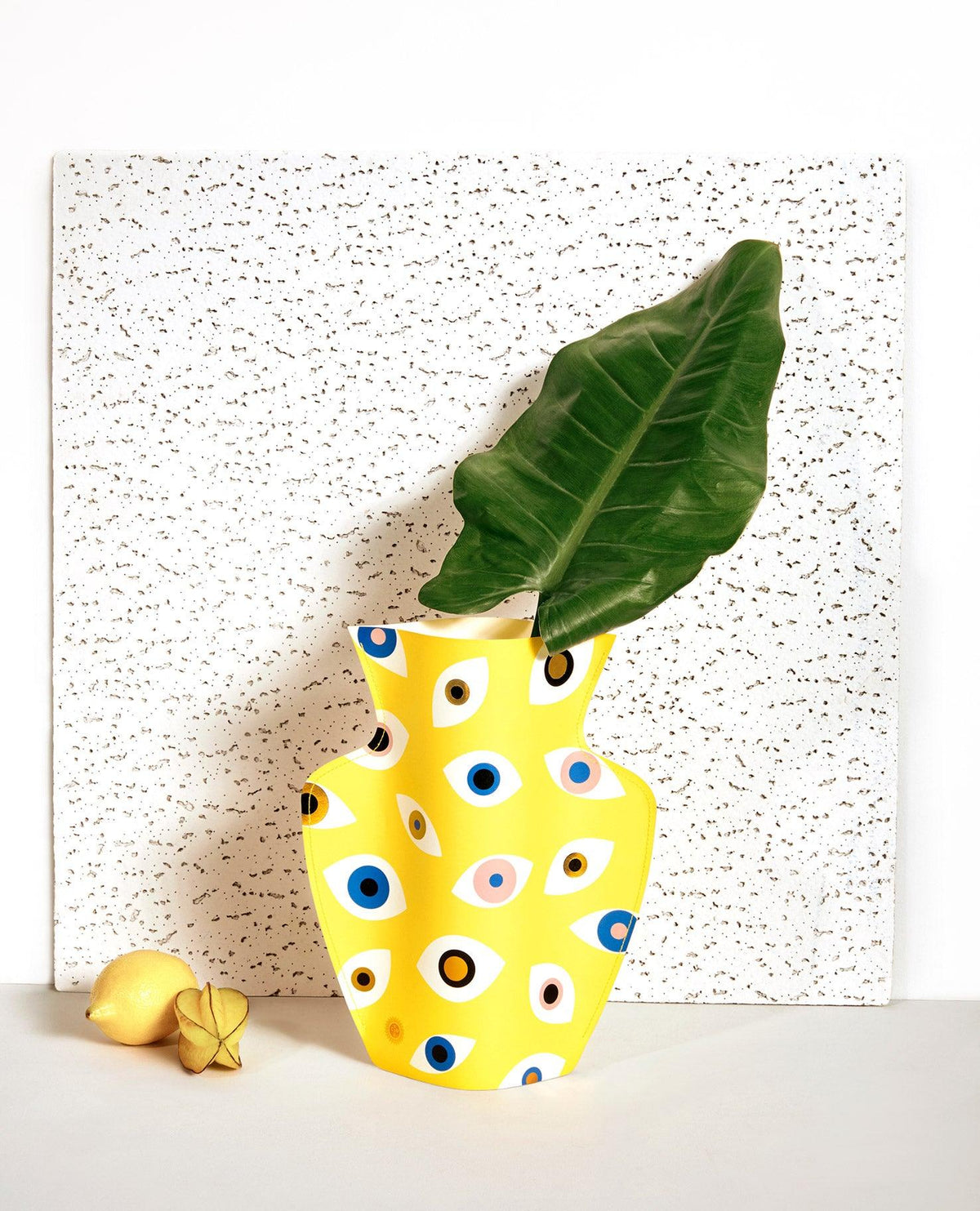 OCTAEVO Nazar Paper Vase in yellow with eye patterns holds a large green leaf, with two lemons nearby.