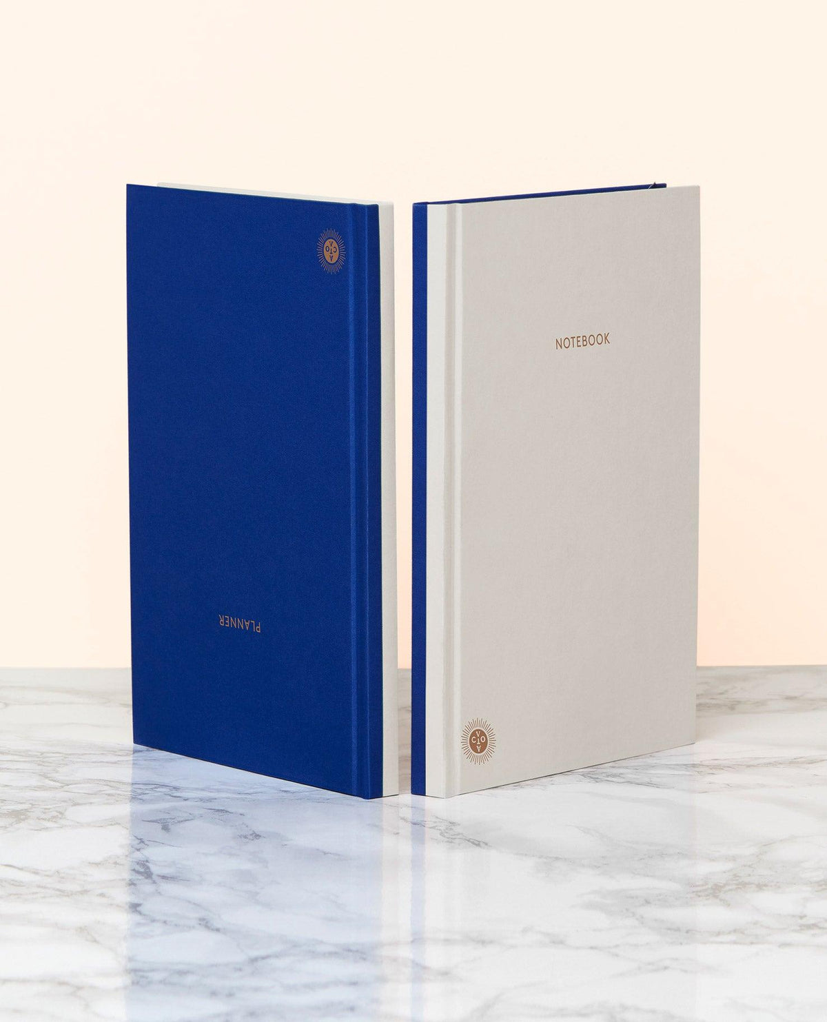 Two OCTAEVO Notebook Planners, blue and grey with gold text, lie on marble; both feature lay-flat hardcover binding.