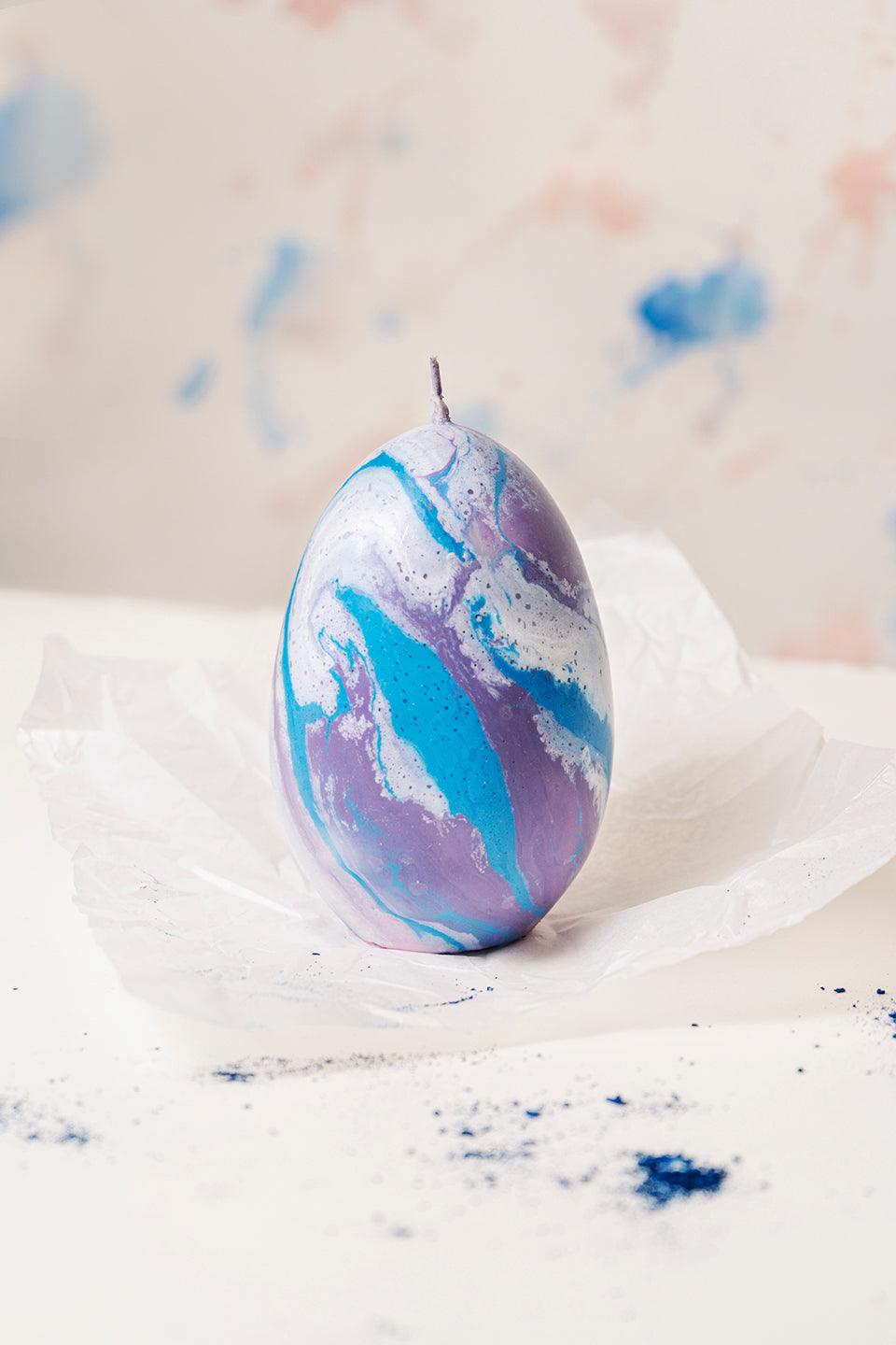 The Olimpo Marble Candle by Octaevo, handmade in Italy, rests on crumpled white paper with blue specks nearby.