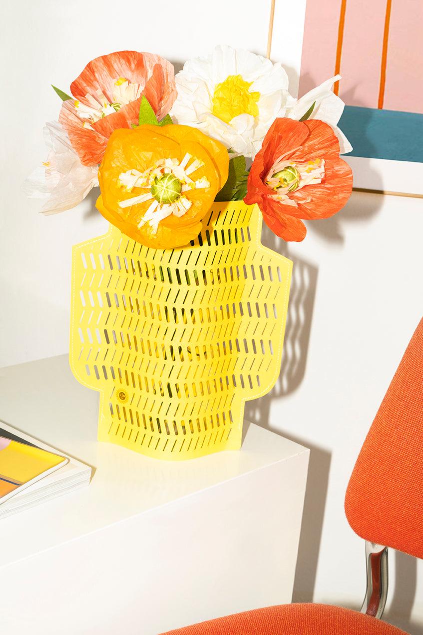 Le bouquet de fleurs en papier Sunset OCTAEVO est posé dans un vase jaune sur un bureau blanc, à côté d'une chaise orange.