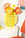 The Octaevo Paper Flower Bouquet Sunset in a yellow geometric vase sits on a white table by an orange chair and wall art.