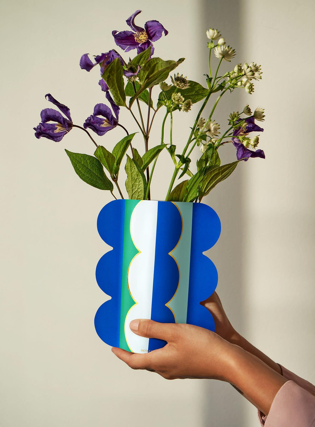 Hands hold an OCTAEVO Riviera Wave Paper Vase with wavy edges, filled with purple and white flowers.