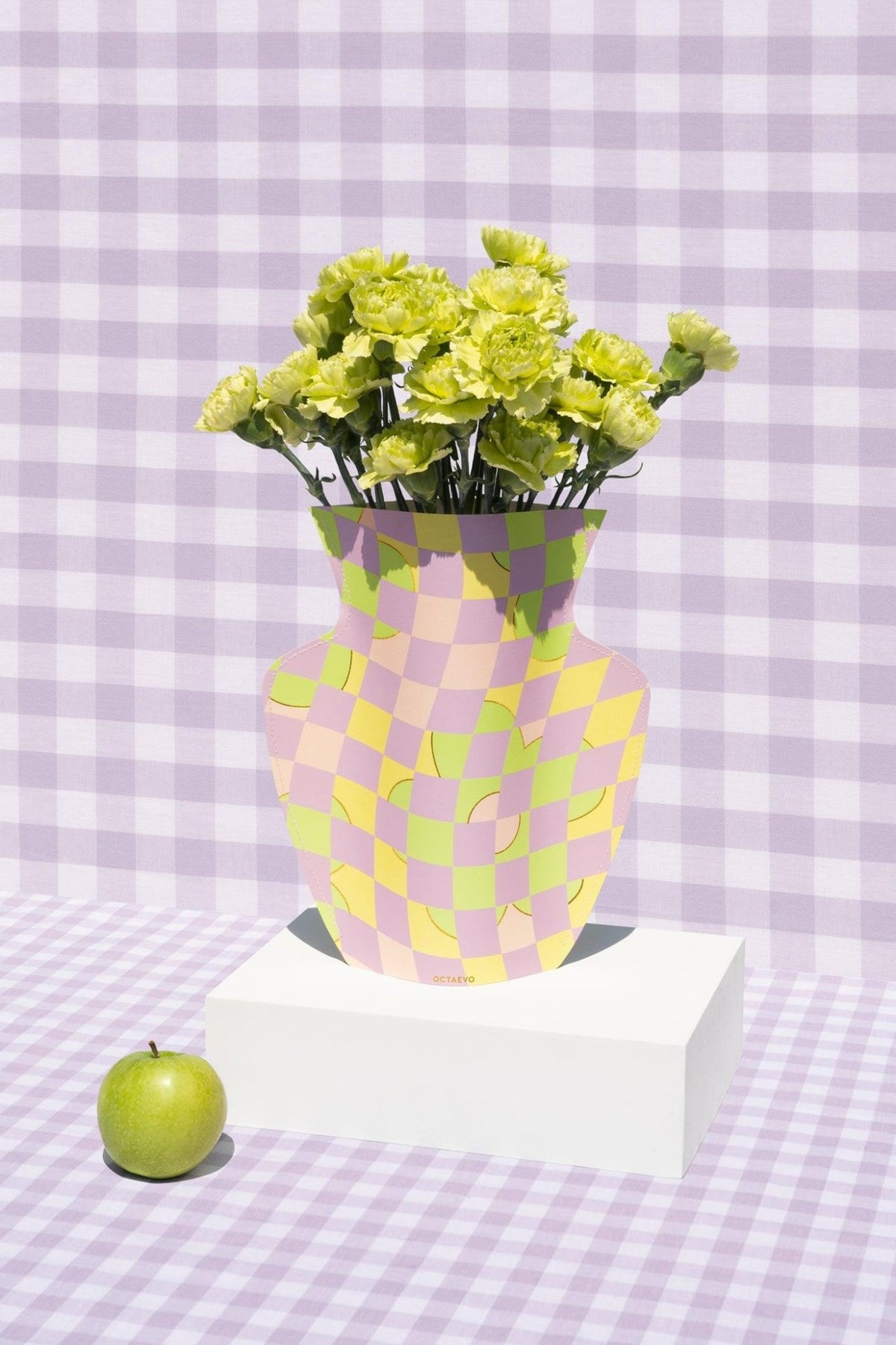 A green apple sits by an Octaevo Picnic Paper Vase with yellow flowers on a white block, against a purple checkered background.