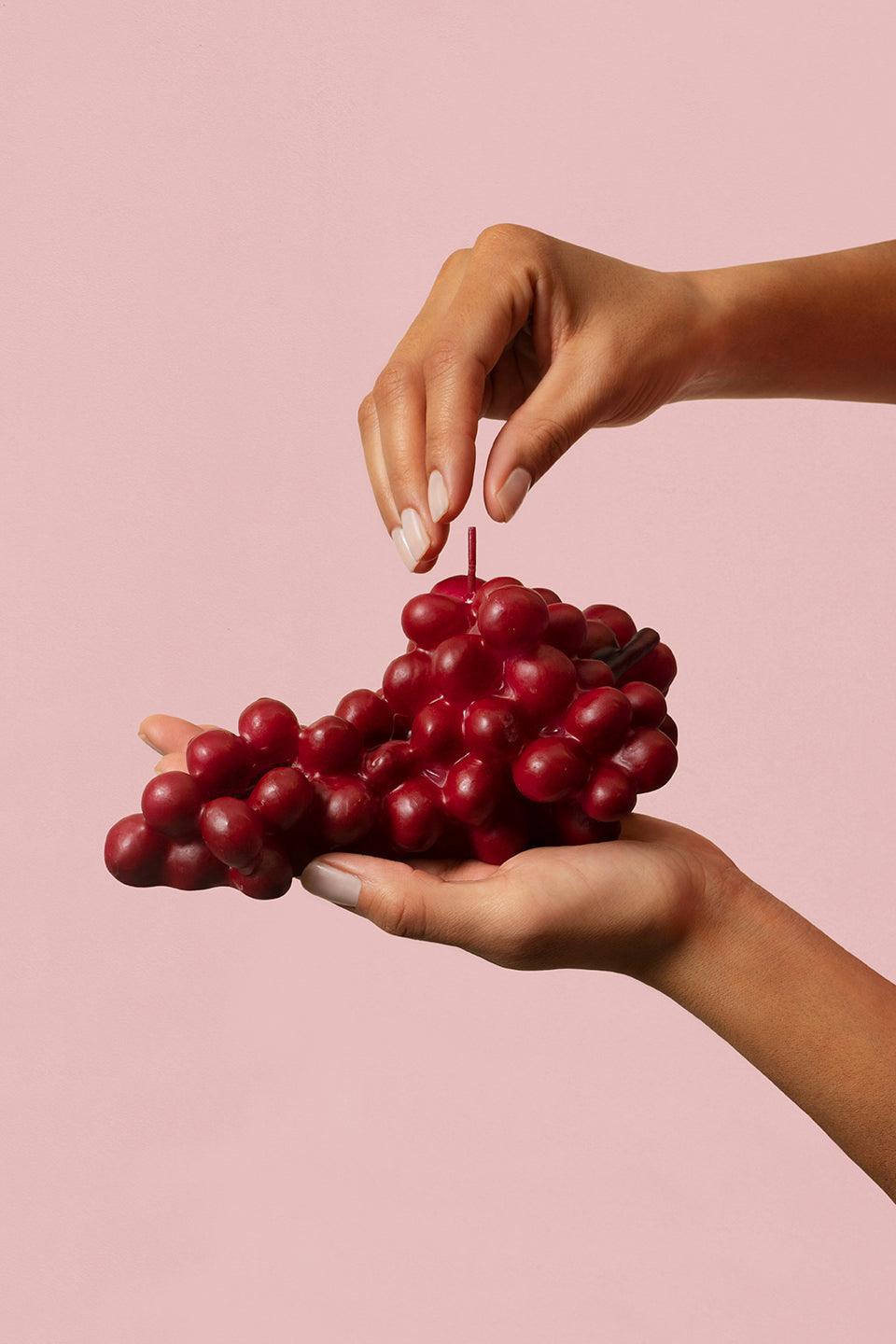 Two hands hold the Octaevo Red Grape Candle against a pink background, an eye-catching decor piece.