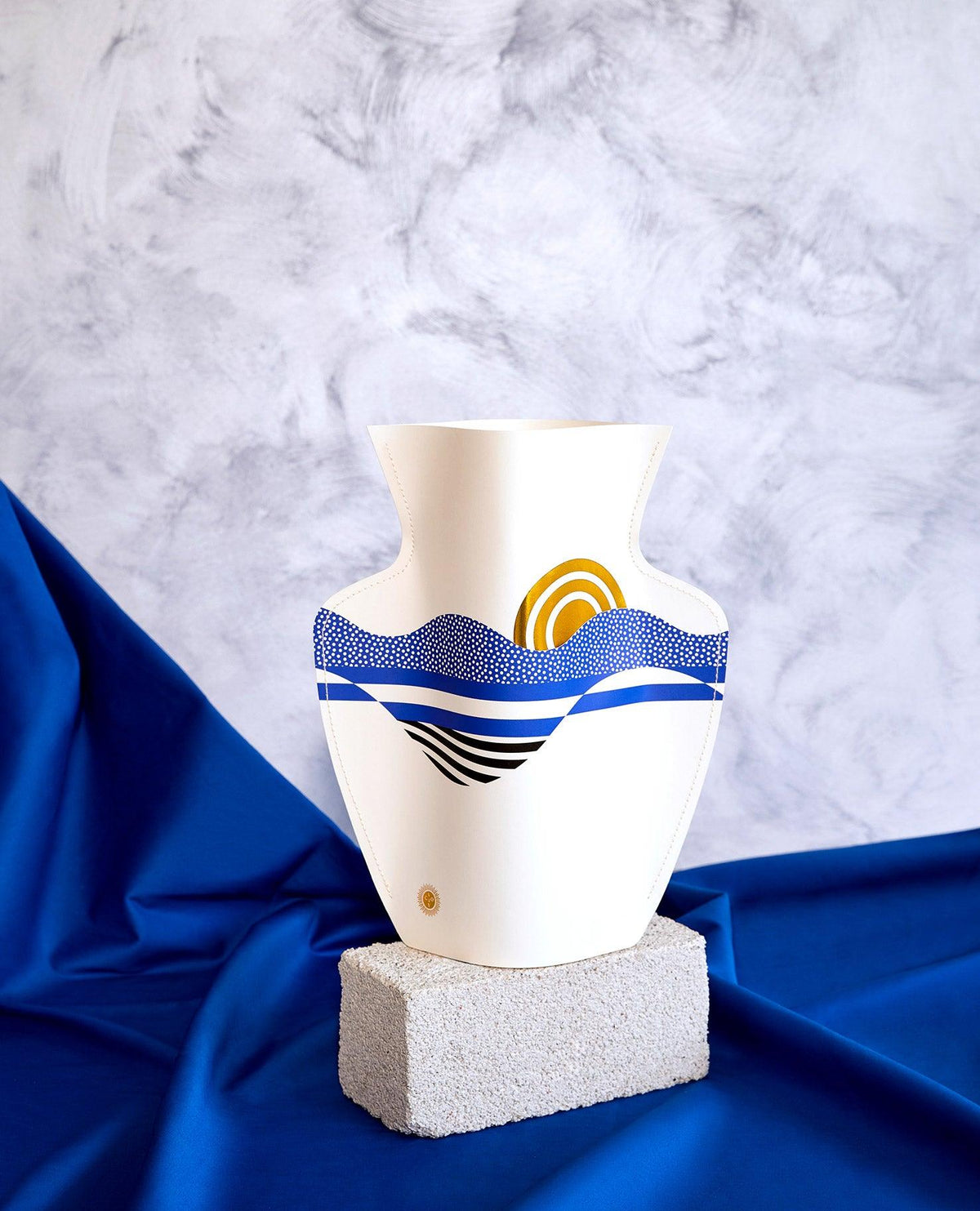 Le vase en papier Salina OCTAEVO arbore des vagues bleues et dorées, posé sur un bloc gris avec un tissu bleu, devant un fond marbré.