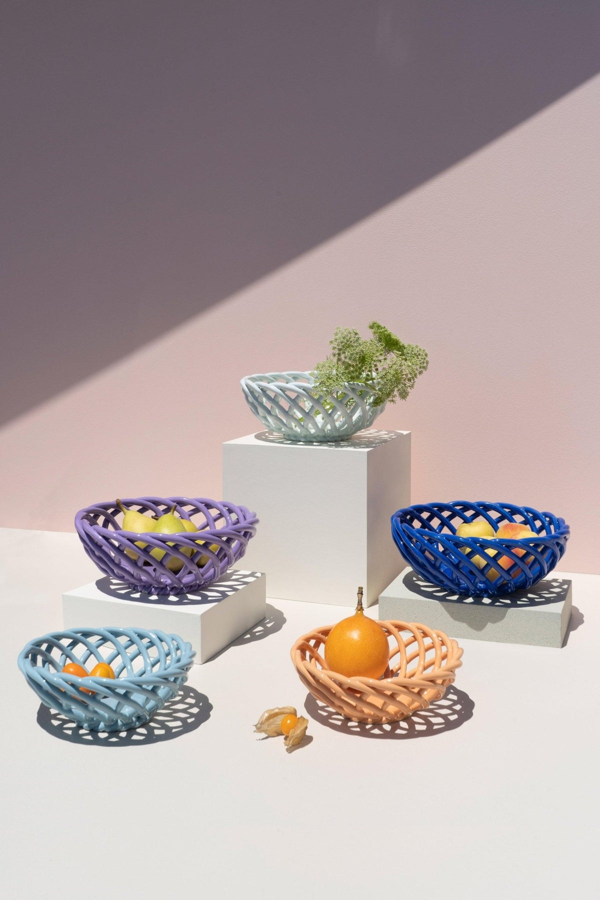 A Sicilia Ceramic Basket by OCTAEVO with fruit and greenery sits as a centerpiece on white blocks and a pink background.