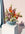 The light blue OCTAEVO Sicilia Ceramic Basket holds a vibrant bouquet on a white pedestal against a pink wall.