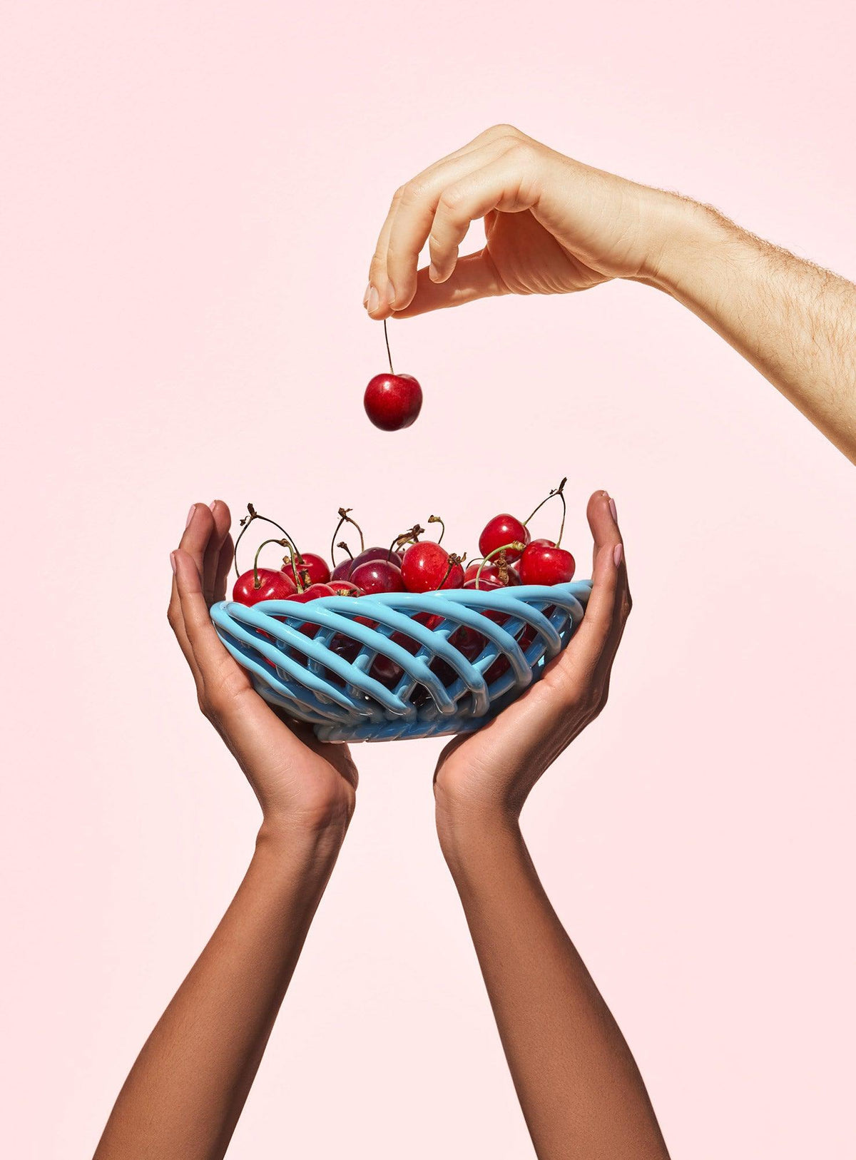 Two hands hold OCTAEVO's Sicilia Ceramic Basket — Small in blue as a cherry is lifted, set against a pink background.