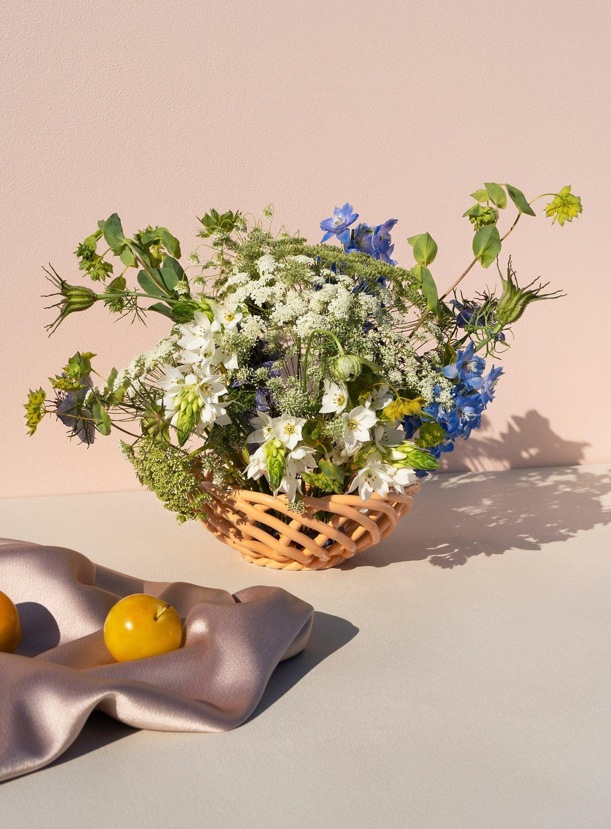 The Sicilia Ceramic Basket — Small by OCTAEVO holds wildflowers beside two yellow fruits on a soft cloth.