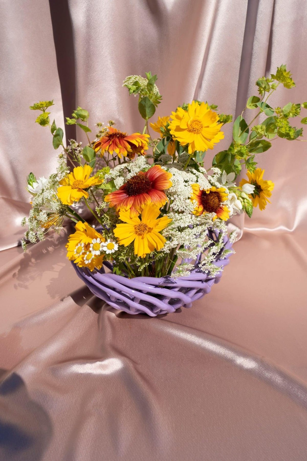 A bouquet of bright wildflowers sits in OCTAEVO’s Sicilia Ceramic Basket — Purple, against metallic pink fabric.
