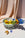 The OCTAEVO Sicilia Ceramic Basket with yellow fruits and blue flowers sits on a table with peach fabric behind.