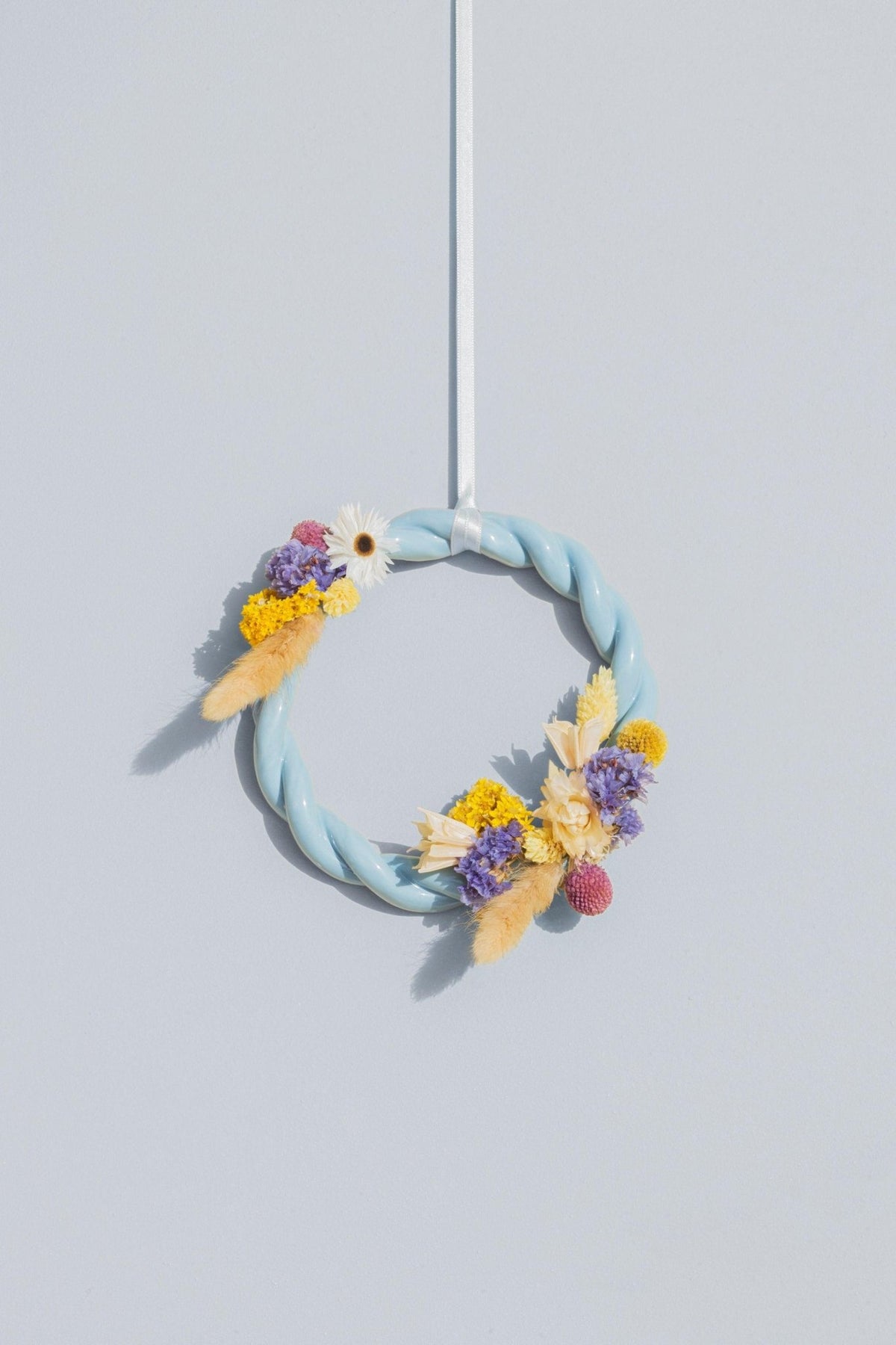 The light blue OCTAEVO Sicilia Ceramic Trivet is displayed against a light gray wall, hanging by a white ribbon.