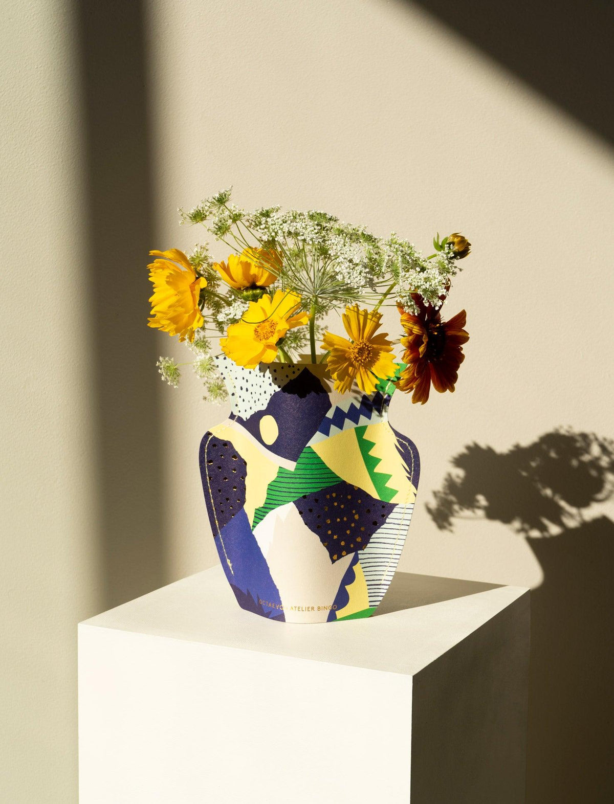 The OCTAEVO Stromboli Mini Paper Vase features a colorful geometric design and holds cheerful yellow and white flowers.