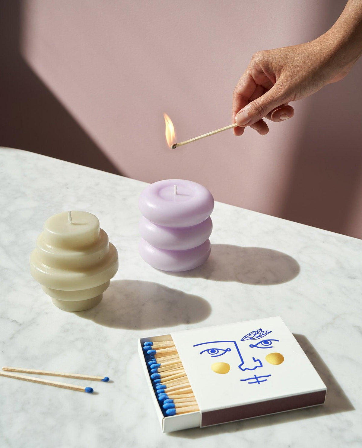 A hand lights a match near OCTAEVO's Templo Candle in Pale Lilac and a matchbox on a marble surface.