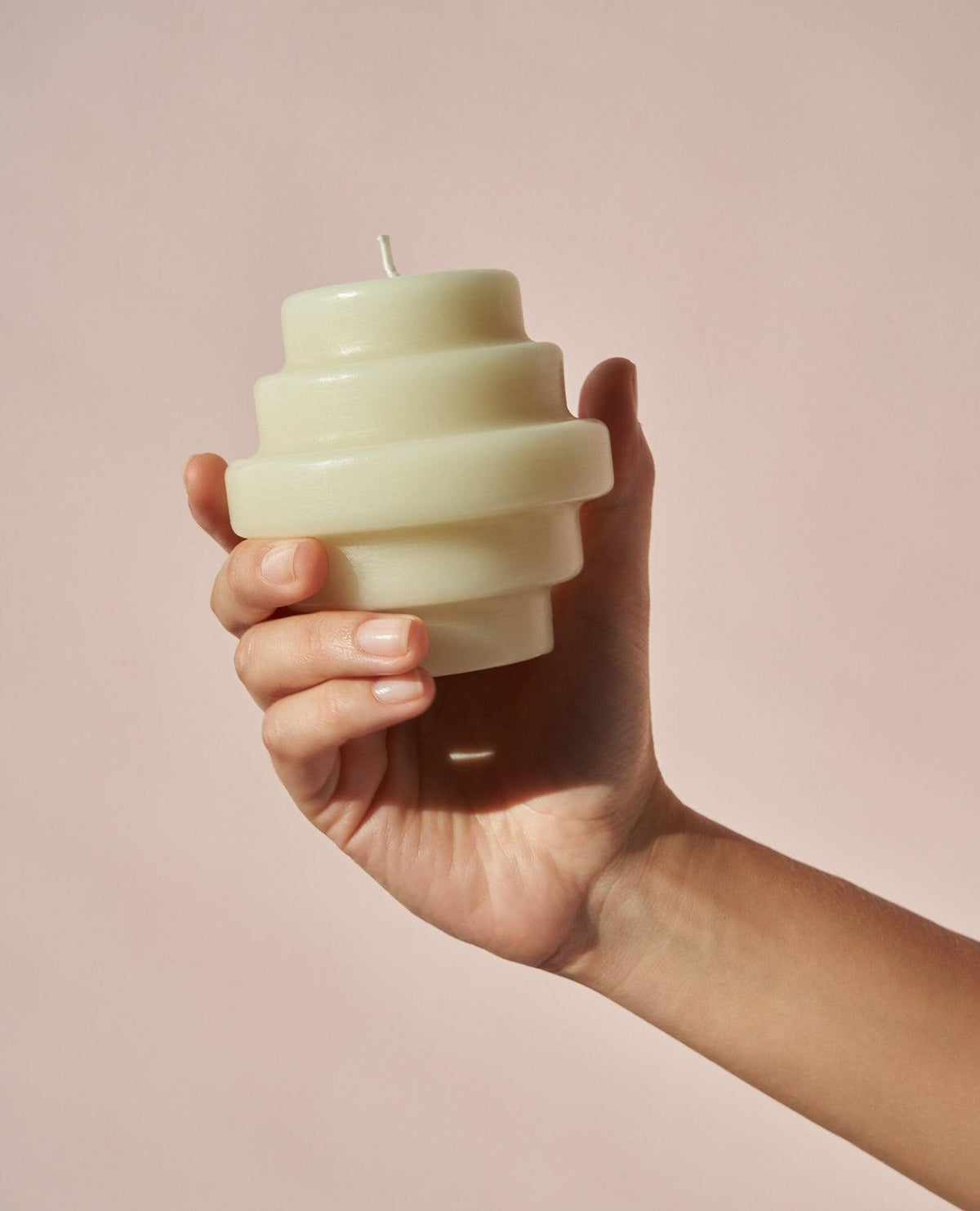 A hand holds the OCTAEVO Templo Candle — Ash Green, its spiral shape glowing softly against a gentle pink background.