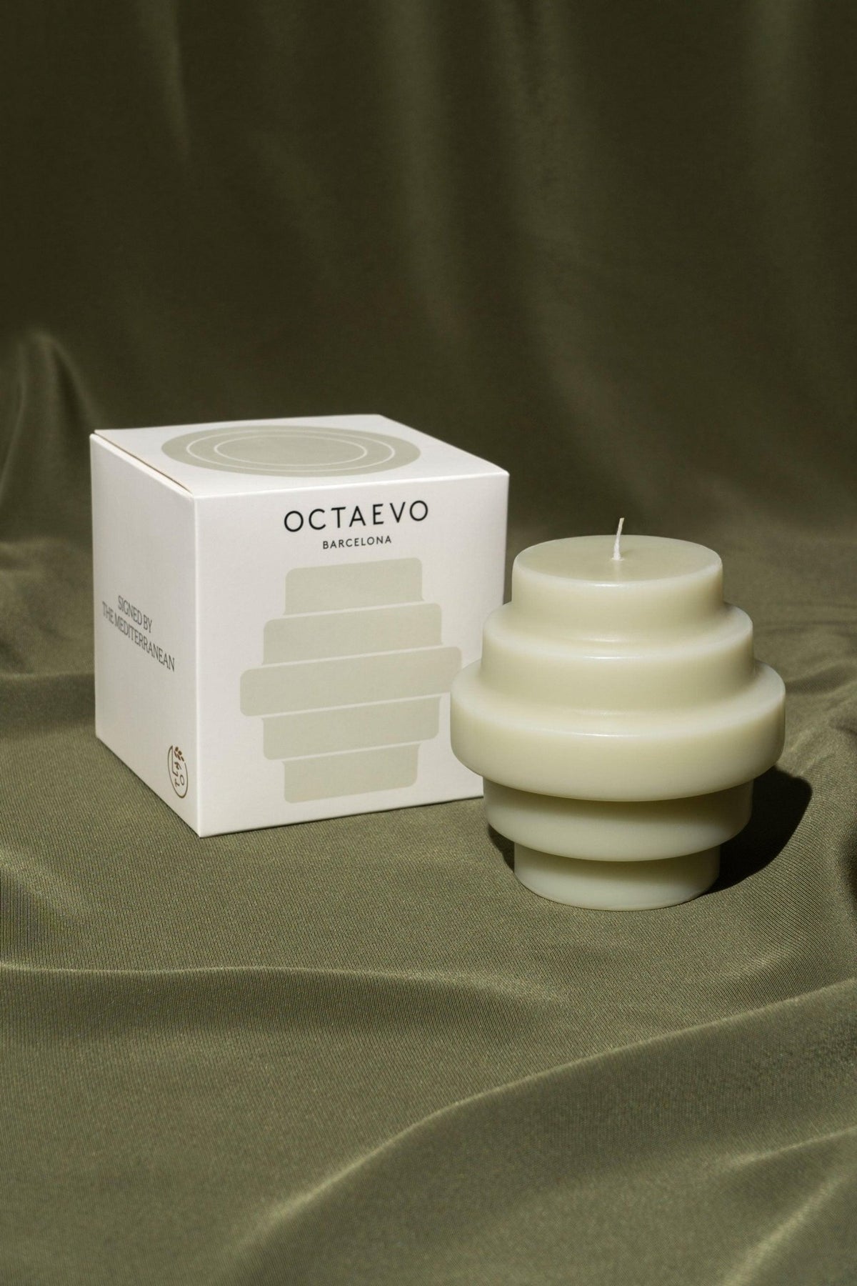 The Templo Candle — Ash Green by OCTAEVO rests in its box on olive-green satin—a striking decorative accent.