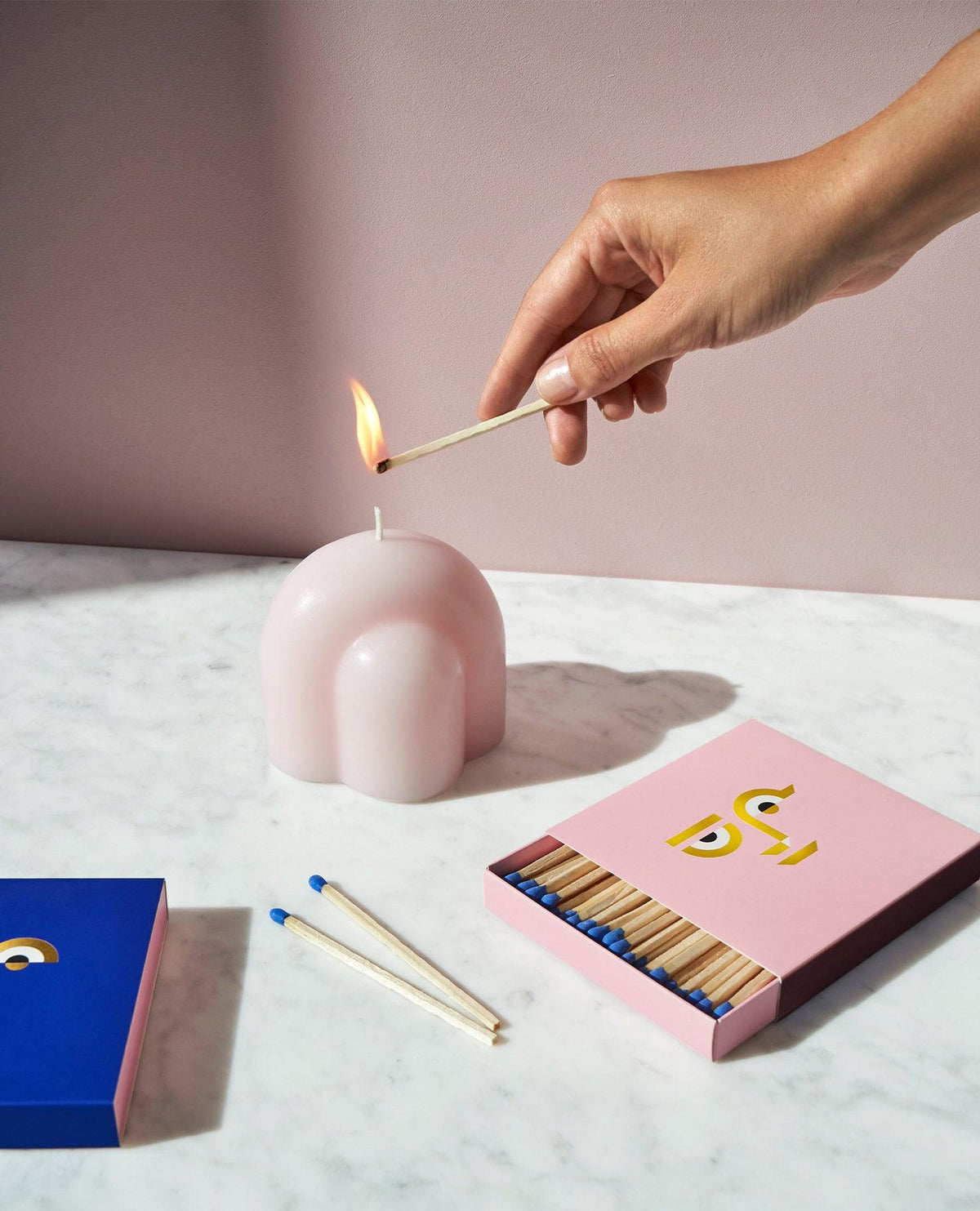 A hand lights OCTAEVO's Templo Candle — Pale Rose on marble with matchboxes—a sculptural, dome-shaped wax art piece.
