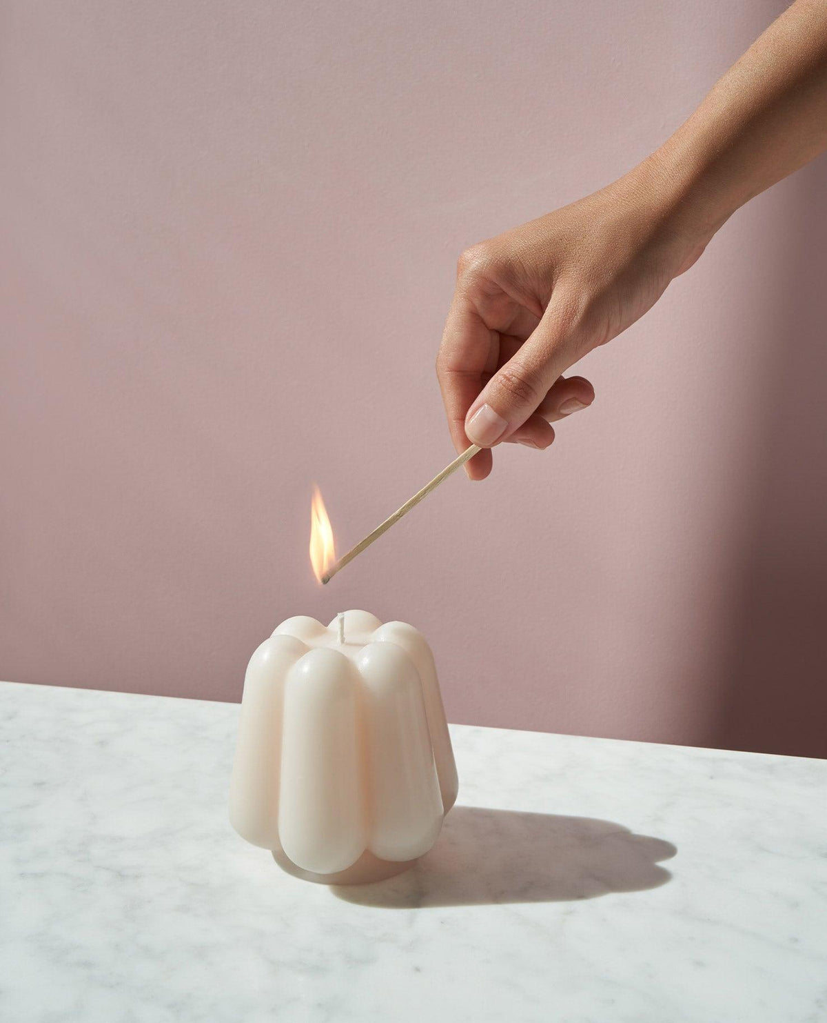 A hand lights OCTAEVO’s Templo Candle — Ivory on marble, set against a pink background.