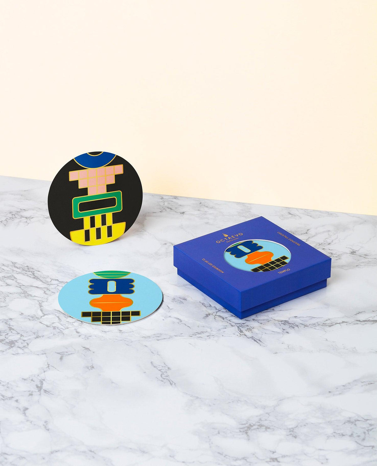 Douze dessous de verre Templo OCTAEVO à visages abstraits sont posés à côté d'une boîte cadeau bleue, sur une surface en marbre.