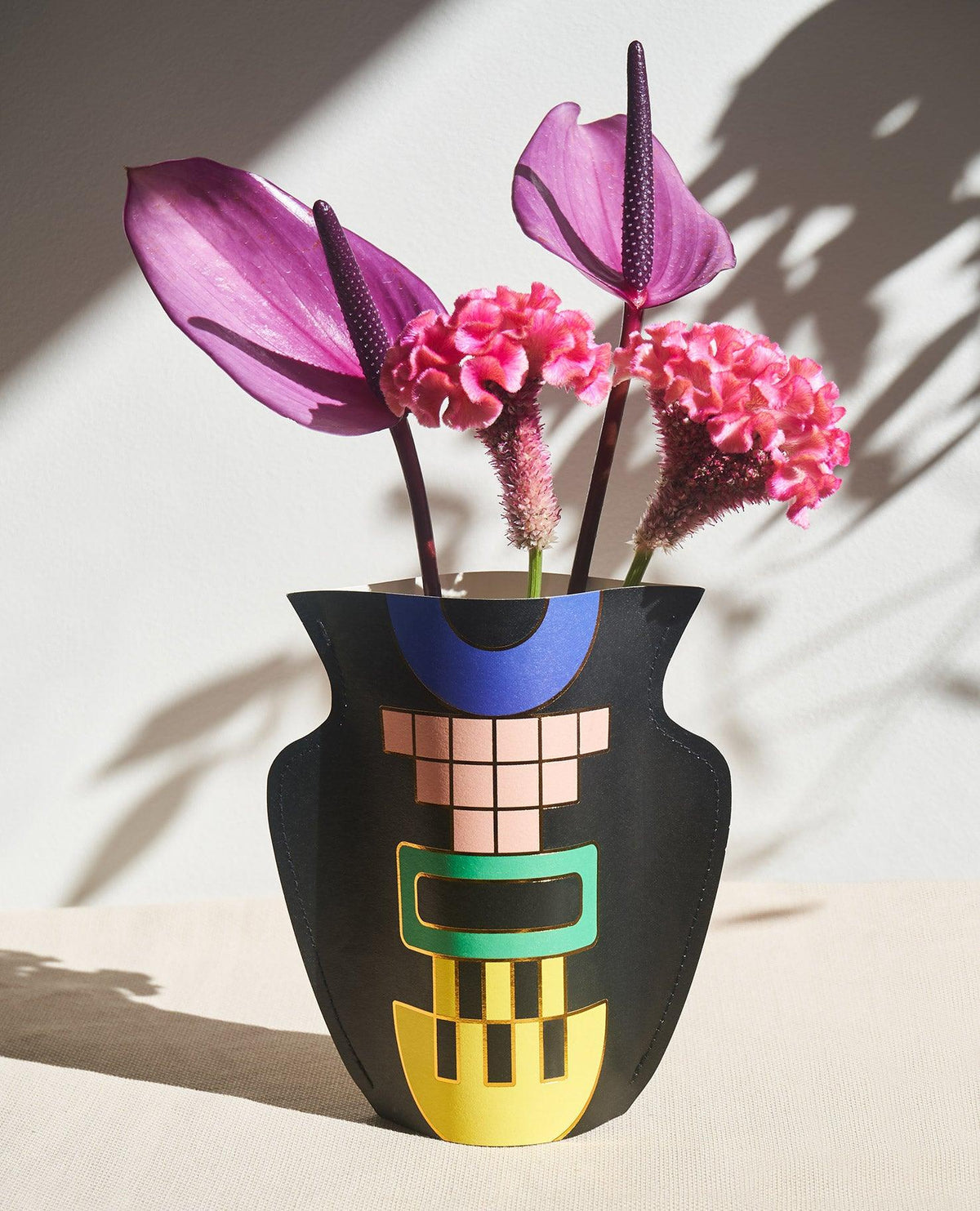 Black OCTAEVO Templo Paper Vase features a colorful geometric design, shown with pink and purple flowers in sunlight.