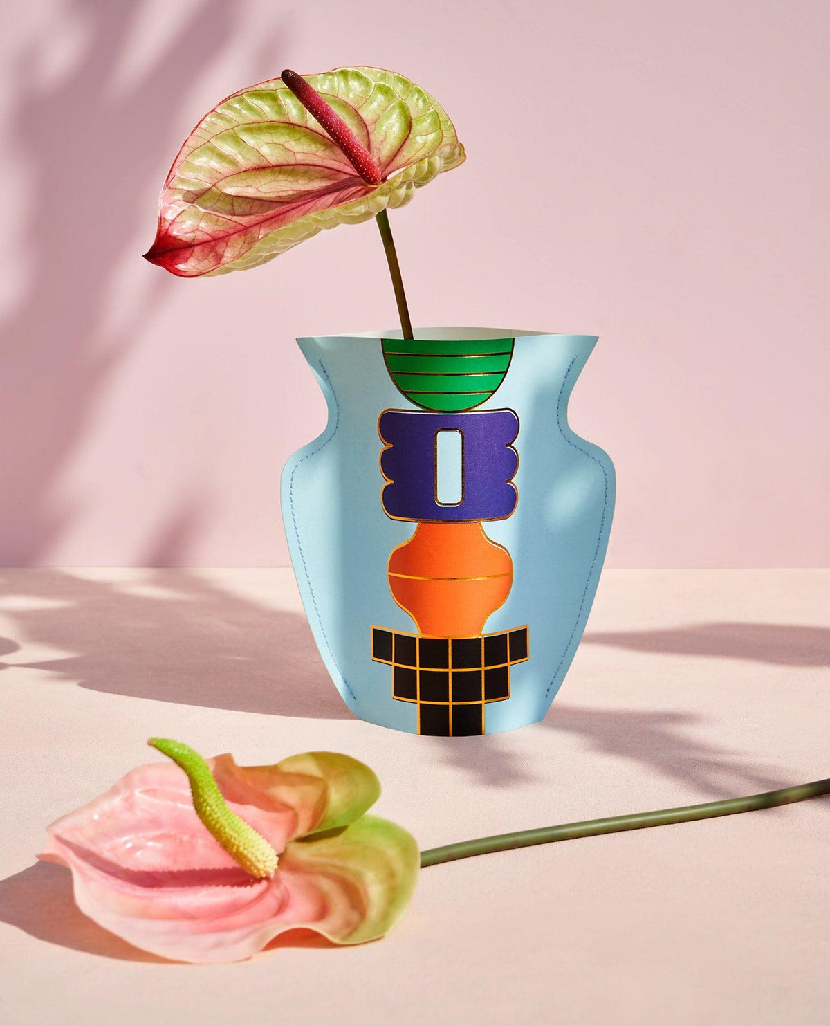 The light blue OCTAEVO Templo Paper Vase with bold geometric patterns holds a pink anthurium; another flower lies nearby.