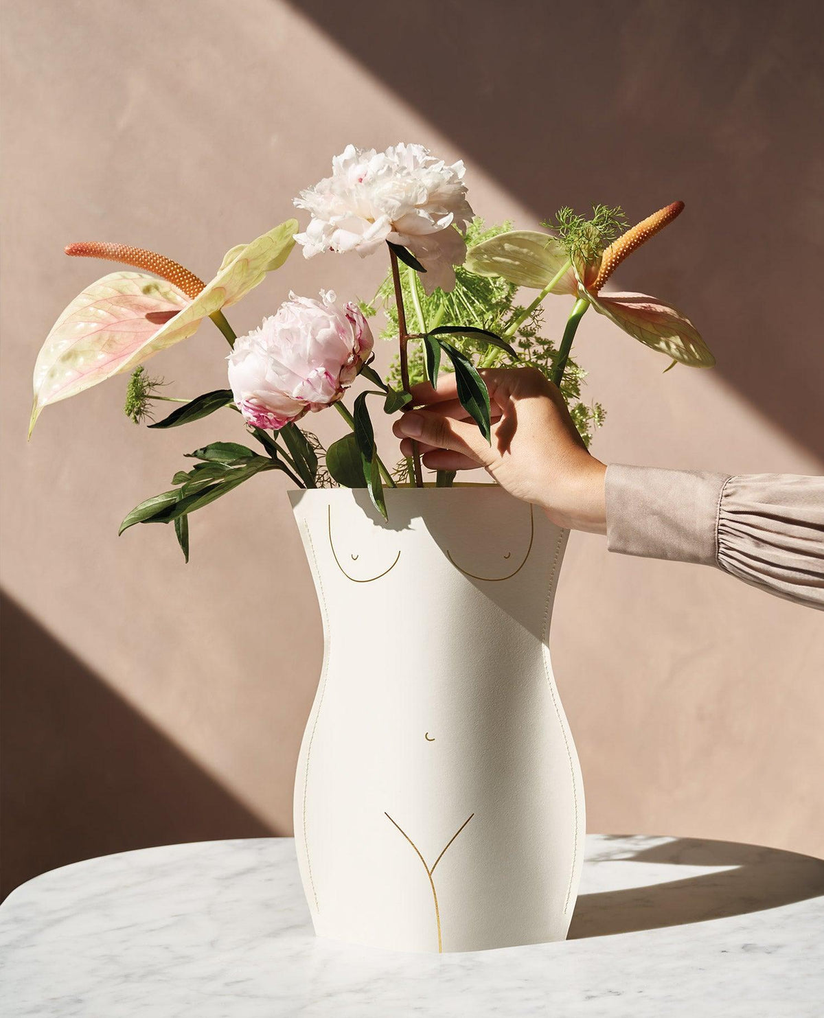 A hand arranges flowers in the OCTAEVO Venus Paper Vase, shaped like a female torso, on a marble table.